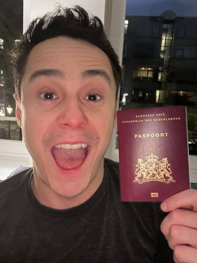 Homem branco de cabelos escuros sorrindo e segurando um passaporte holandês.